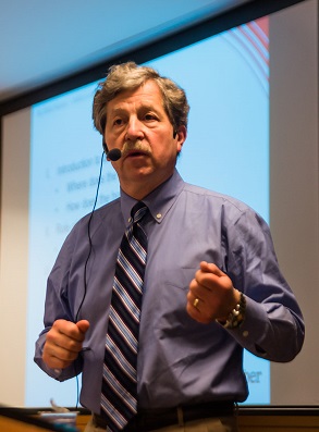 Charles A Nelson wears an ear piece microphone and stands in front of a powerpoint as he presents.