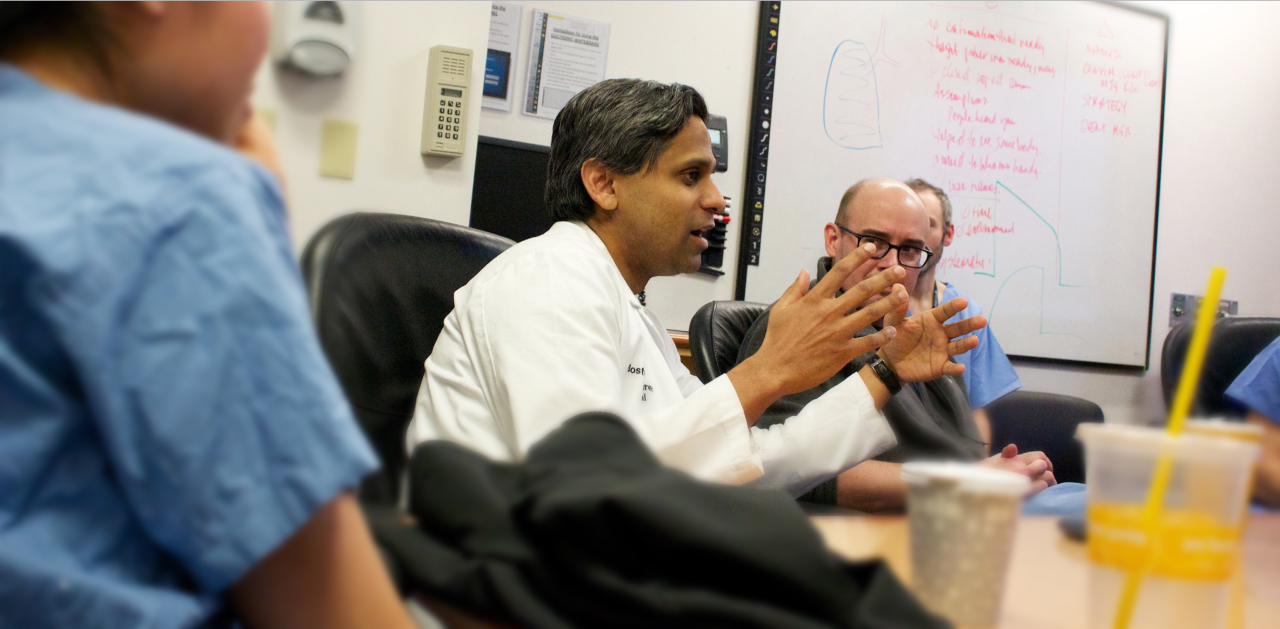 A man with a white coat sits in the middle of a table while gesturing with his hands.