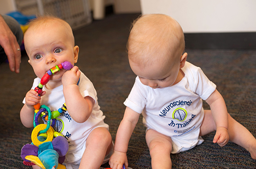 Two babies in white onesies sit on a grey carpet and play with plastic teethers