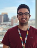 A man, Lucas Francisco, wears glasses and a red shirt as he stands outside and smiles at the camera.