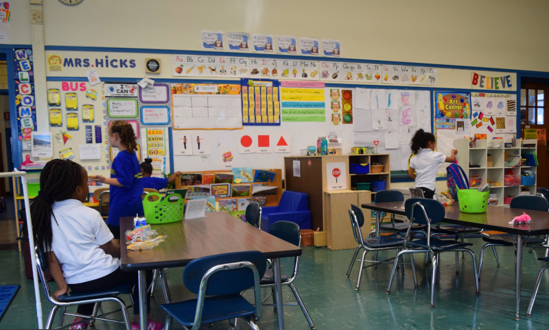 Children stand and sit at desks in a classroom.